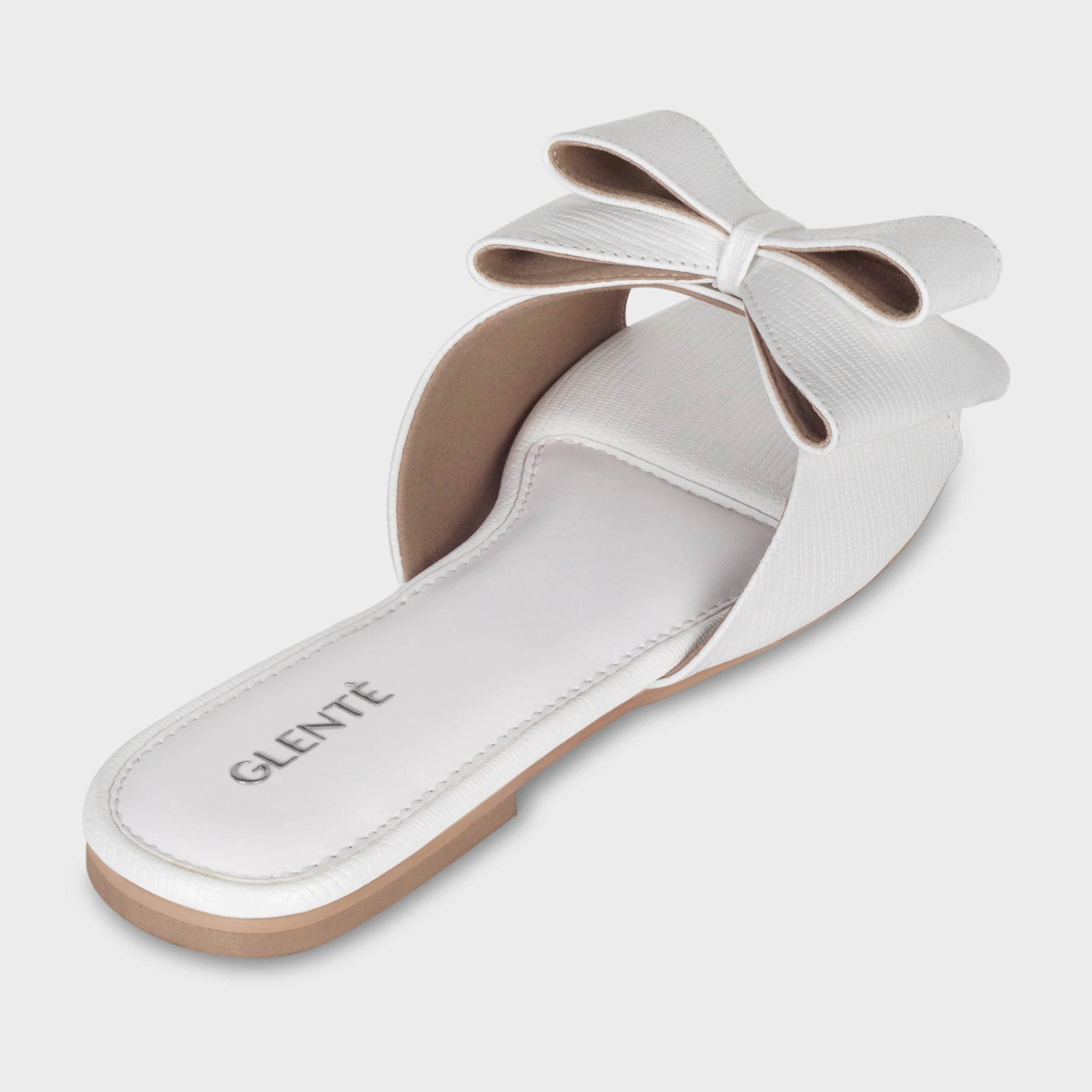 Rear angled view of Alveria white flat slides showing the padded white insole with silver Glentè branding, raised bow silhouette above the strap, and lizard-textured trim
