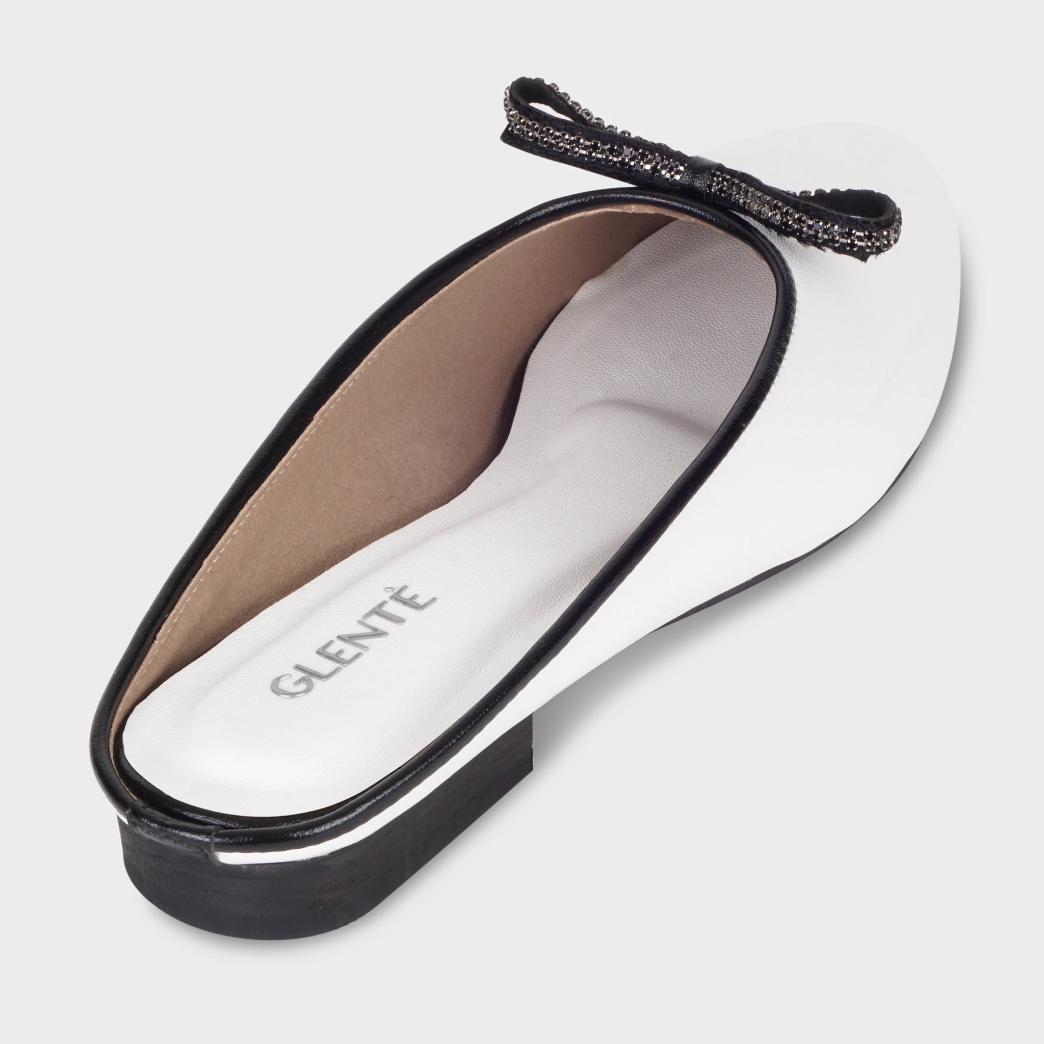 Rear angled view of Auralin white mules showing the cushioned interior with silver Glentè branding, black crystal bow at the vamp, and contrast black heel and sole edge