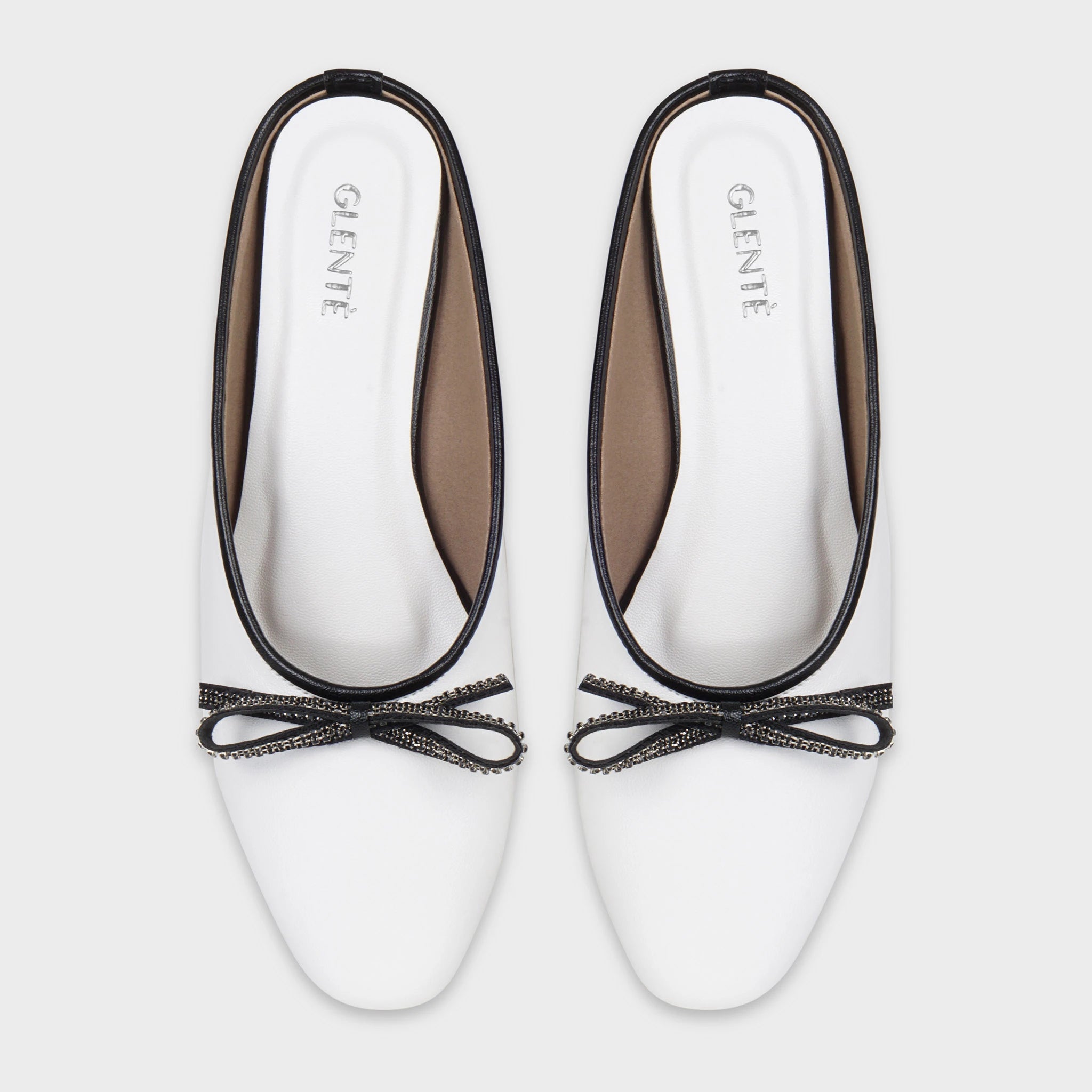 Top-down view of Auralin white flat mules as a pair, showing the symmetrical black rhinestone bow accents, clean round-toe shape, contrast black piping, and silver Glentè branding on white insoles