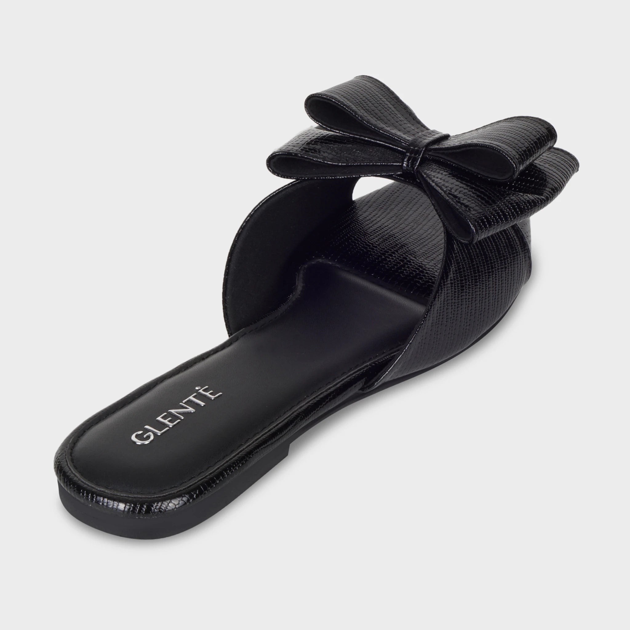 Rear angled view of Alveria black flat slides showing the cushioned black insole with silver Glentè branding, sculptural bow rising above the strap, and lizard-textured edging
