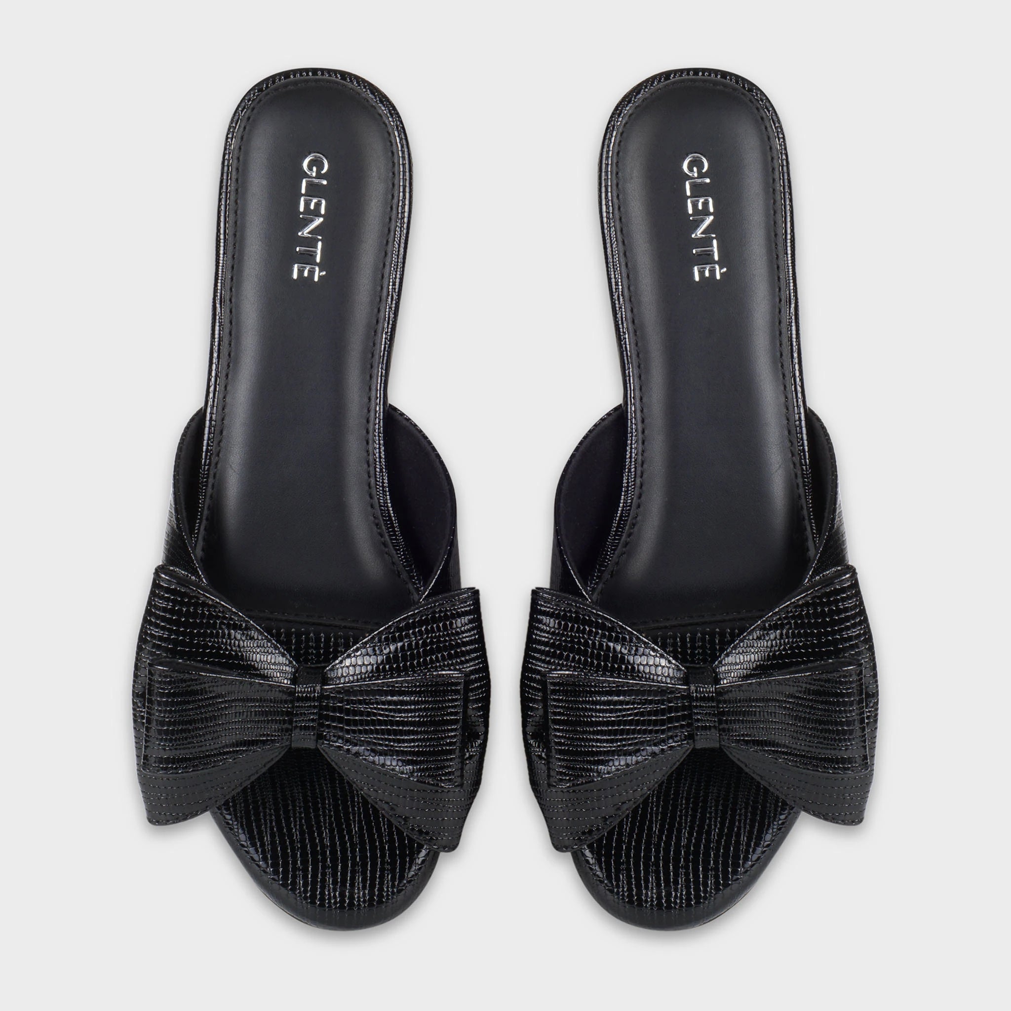 Top-down view of Alveria black lizard-embossed flat slides as a pair, showing the symmetrical double bow accents, wide strap design, and silver Glentè branding on black insoles