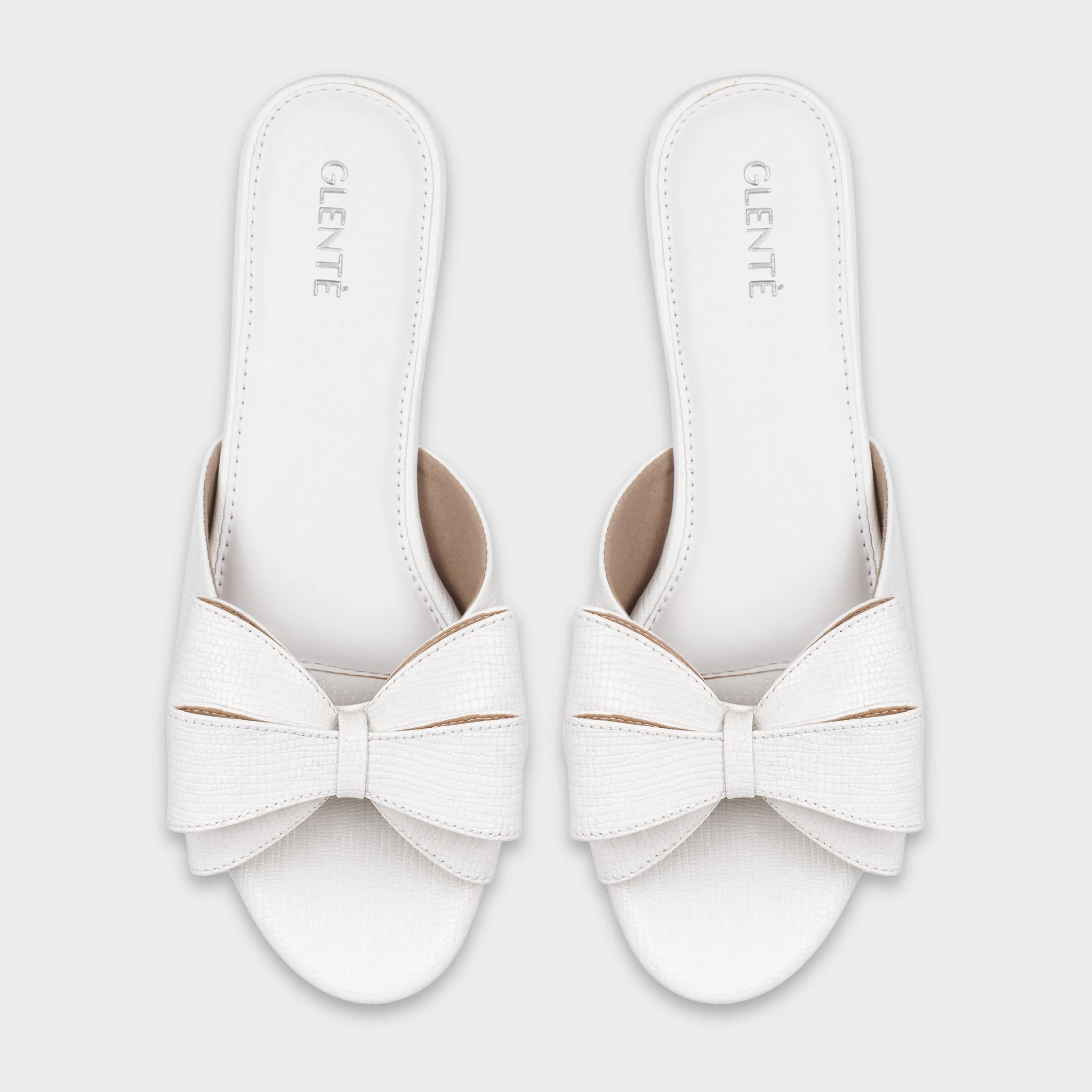 Top-down view of Alveria white lizard-embossed flat slides as a pair, showing the matching double bow accents, wide single-strap design, and silver Glentè branding on white insoles