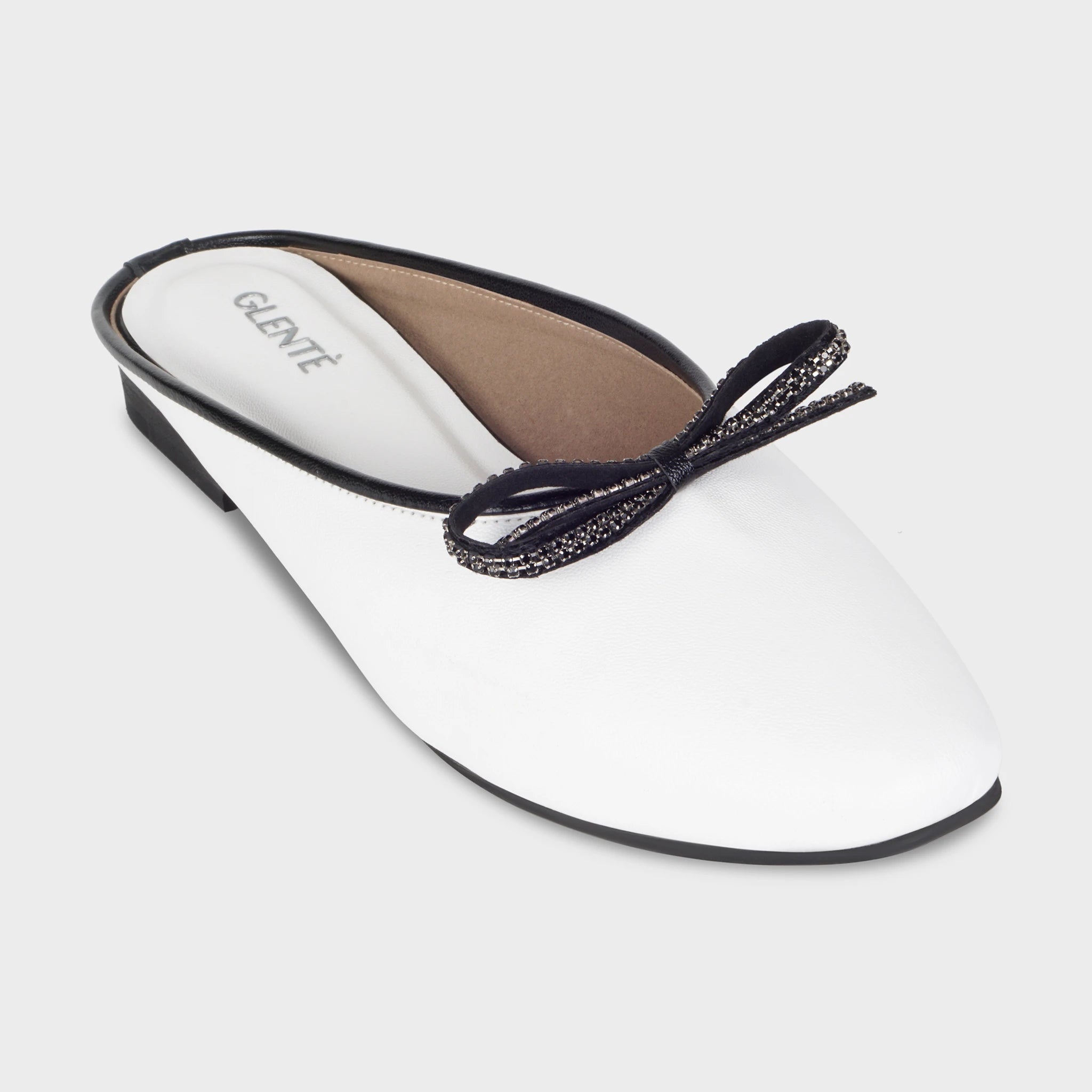 Front angled view of Auralin white flat mules highlighting the round toe, black rhinestone-studded bow with trailing loops, and silver Glentè branding on the white insole