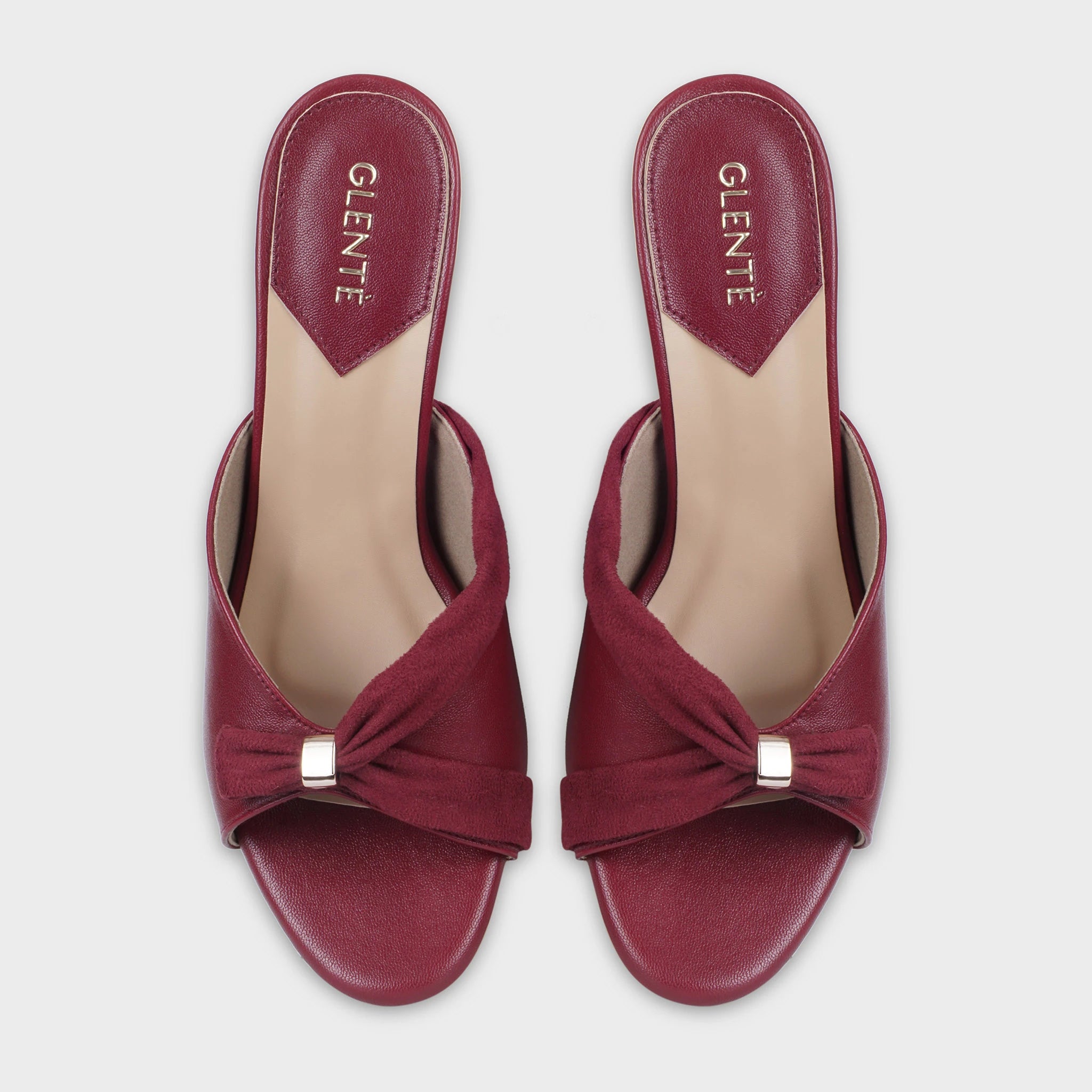 Top-down view of Solviane cherry red heeled mules as a pair, showing the symmetrical suede twisted knot design with gold hardware, open toe cut, and gold Glentè branding on cream insoles