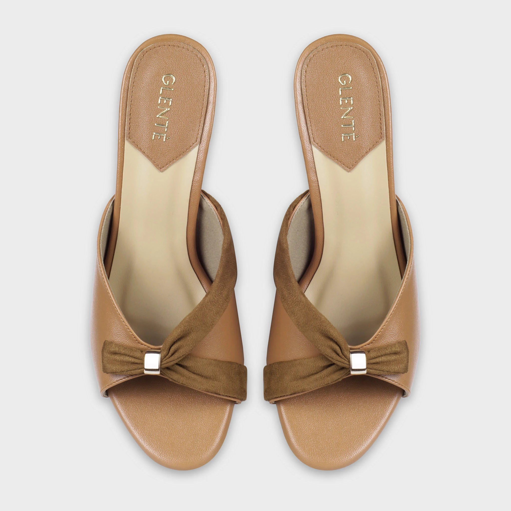 Top-down view of Solviane tan heeled mules as a pair, showing the symmetrical brown suede twisted knot design with gold hardware, open toe cut, and gold Glentè branding on cream insoles
